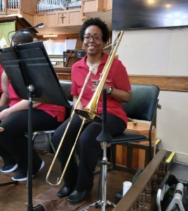 Natalya sits holding her trombone. She is wearing a red JOY Band shirt. 